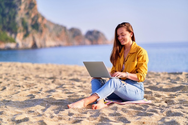 Young happy joyful carefree smiling satisfied millennial freelancer girl using laptop on sand beach by the sea Dream office work concept