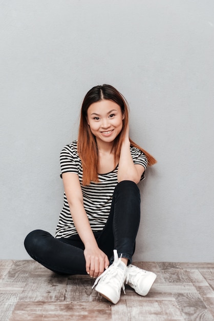 Young happy lady sitting on floor over grey wall.