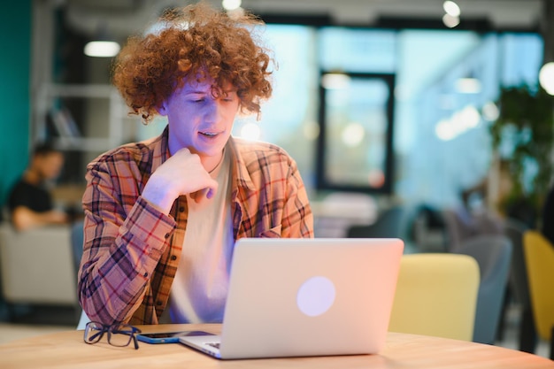 Young happy male freelancer in casual clothes sitting in cafe with laptop and using mobile phone