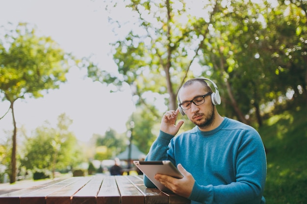 Young happy man, businessman or student in casual blue shirt glasses sitting at table with headphones, tablet pc in city park, listen music, rest outdoors on green nature. Lifestyle leisure concept.