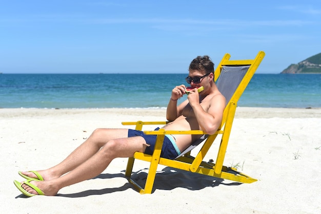 Young happy man eating watermelon sitting on deck chair on a south china sea beach in Vietnam