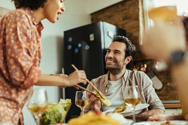 Young happy man sitting at dining table while his African American girlfriend is serving lunch