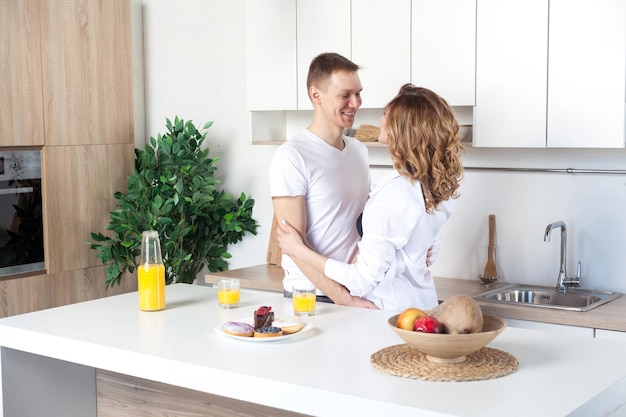 Young happy married couple embraces standing near table in kitchen. Husband hugs his pregnant wife, parents enjoy each other looking to the eyes. Love story, happy people, future parents