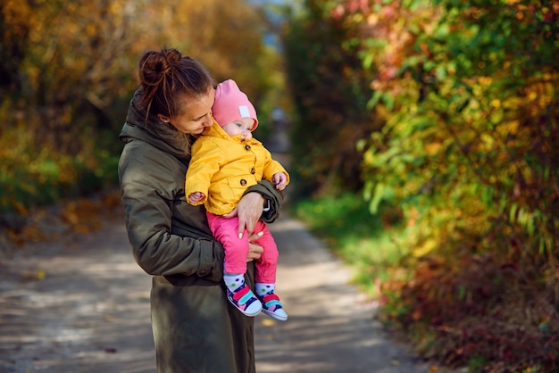 Young happy mother holds her little daughter in a yellow jacket on autumn park. Mother with the child on autumn trees.