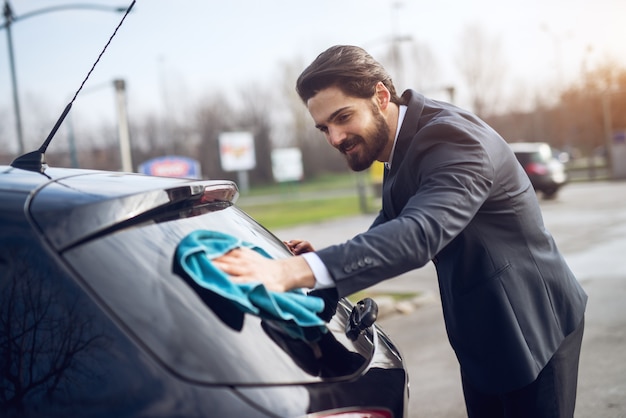 Young happy motivated hardworking young businessman in suit cleaning the rear window of his car with a microfiber cloth at the manual car washing self-service station.