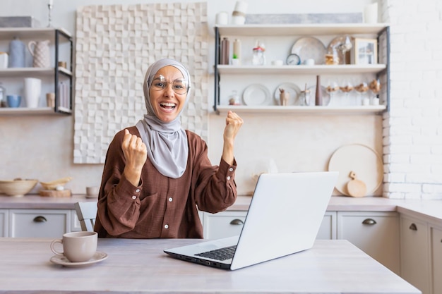 Young happy muslim student woman in hijab sitting at home studying on laptop shows a yes gesture