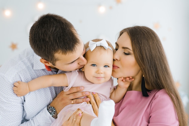 Young happy parents hold their one-year-old daughter in their arms and kiss her on the cheek