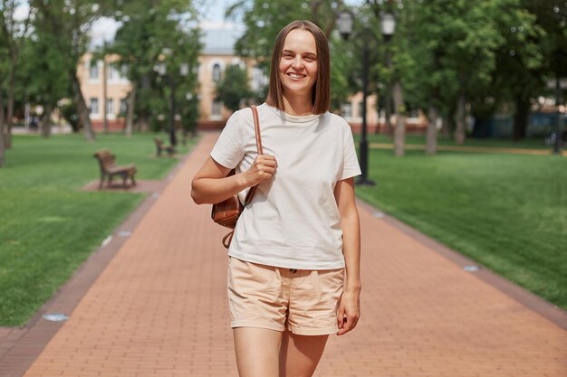 Young happy and pretty brown haired girl wearing white Tshirt and shorts holding bag looking at camera with happy expression walking in city