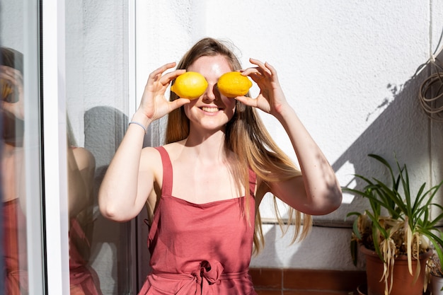 Young happy pretty female in casual summer dress sitting on terrace and covering eyes with ripe lemons