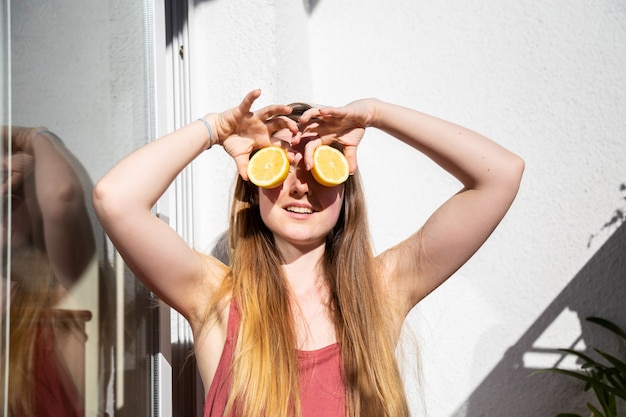 Young happy pretty female in casual summer dress sitting on terrace and covering eyes with ripe lemons