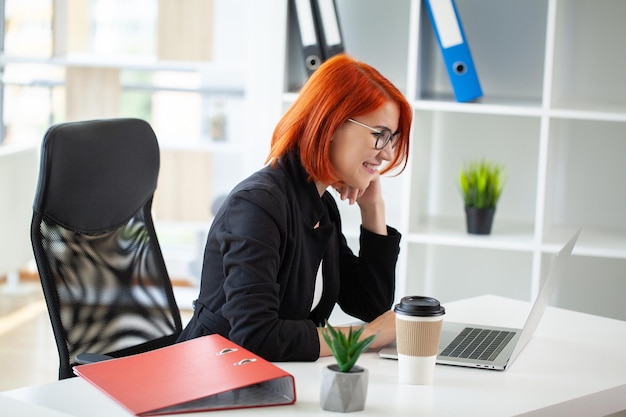 Young happy redhead business woman sit indoors in office using laptop computer