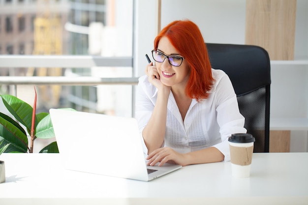 Young happy redhead business woman sit indoors in office using laptop computer