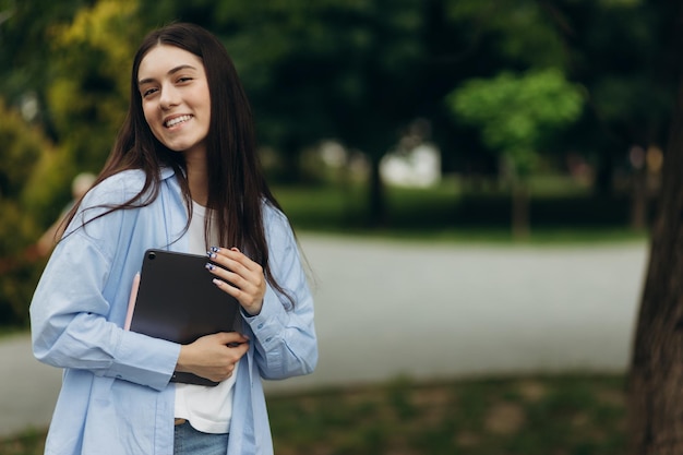 Young happy smiling caucasian young girl business student walking concept in the summer green park outdoor holding tablet Copy space