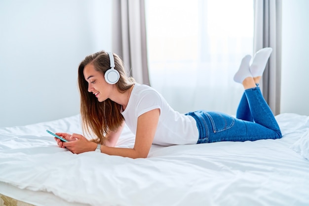 Young happy smiling joyful millennial girl with wireless headphones lying on a bed and using phone for browsing web, social networking, chatting and watching movie online at home