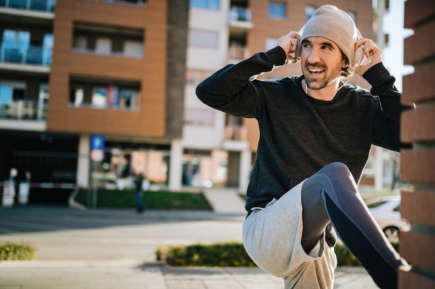 Young happy sportsman adjusting his headphones while stretching and warming up on the street