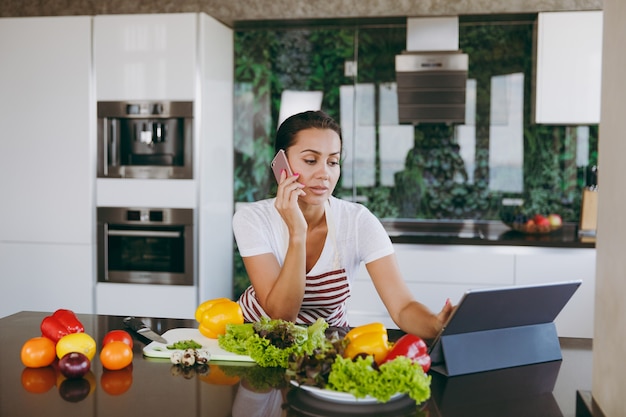 The young happy woman in apron talking on mobile phone and looking at recipe in laptop in the kitchen