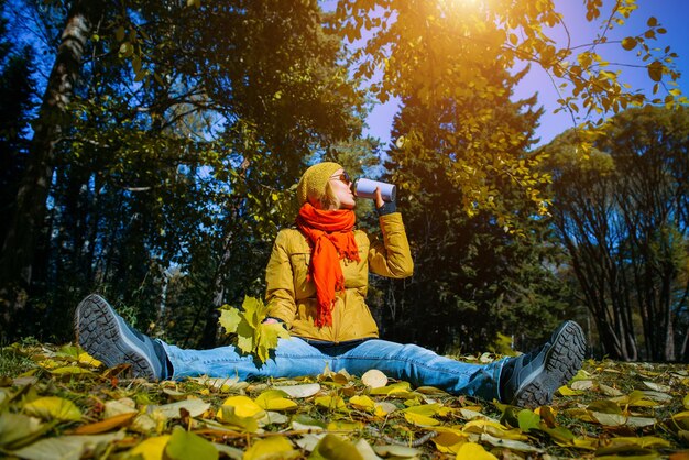 Young happy woman in autumn park on sunny day, drinking coffee or tea, sitting on falling foliage, having fun with yellow leaves. Positive lifestyle concept.