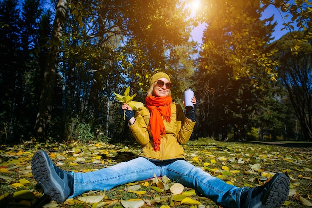 Young happy woman in autumn park on sunny day, drinking coffee or tea, sitting on falling foliage, having fun with yellow leaves. Positive lifestyle concept.