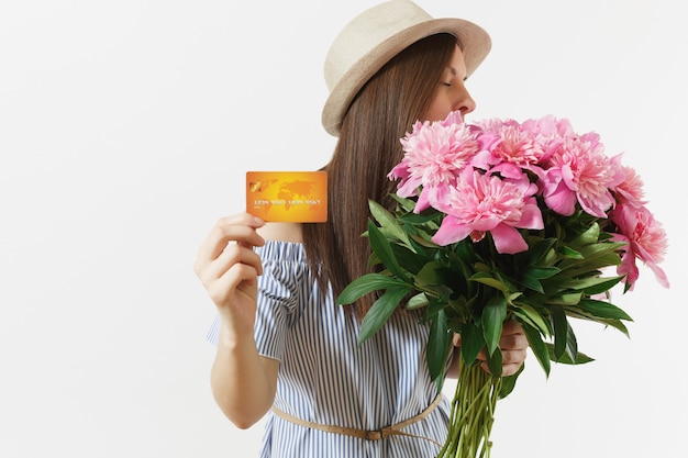 Young happy woman in blue dress, hat holding credit bank card, money, bouquet of beautiful pink peonies flowers isolated on white background. Business, delivery, online shopping concept. Copy space.