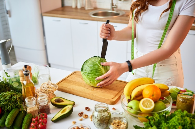 Young happy woman cutting cabbage for making salad in the beautiful kitchen with green fresh ingredients indoors. Healthy food and Dieting concept. Loosing Weight