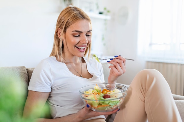 Young and happy woman eating healthy salad sitting on the sofa with green fresh ingredients. Woman eating vegetable salad . Healthy diet