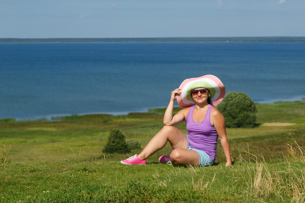 Young happy woman in hat and sunglasses is sitting on a hill in front of a lake.