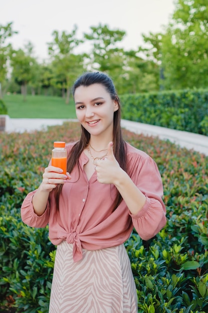 Young happy woman holding bottle with freshly squeezed orange juice Summer day outdoors in the park