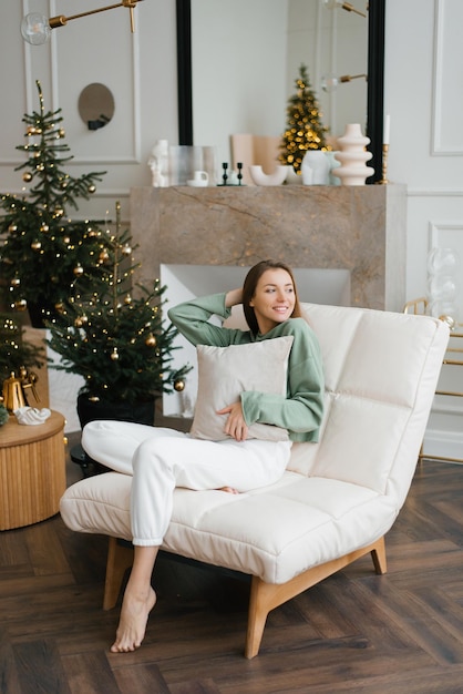 Young happy woman holds a pillow in her hands while sitting in a cozy armchair in the living room decorated for Christmas