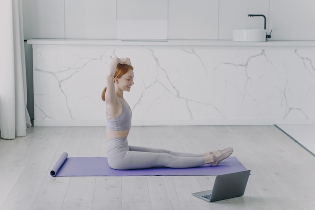 Young happy woman is stretching sitting on floor and doing morning workout in living room