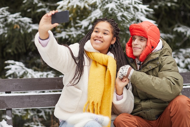 Young happy woman making selfie on her mobile phone with her boyfriend while they sitting on bench in winter