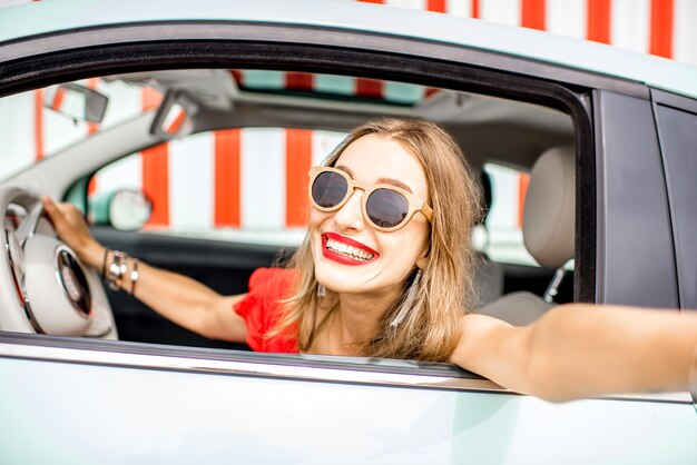 Young and happy woman making selfie photo sitting at the car on the red wall background