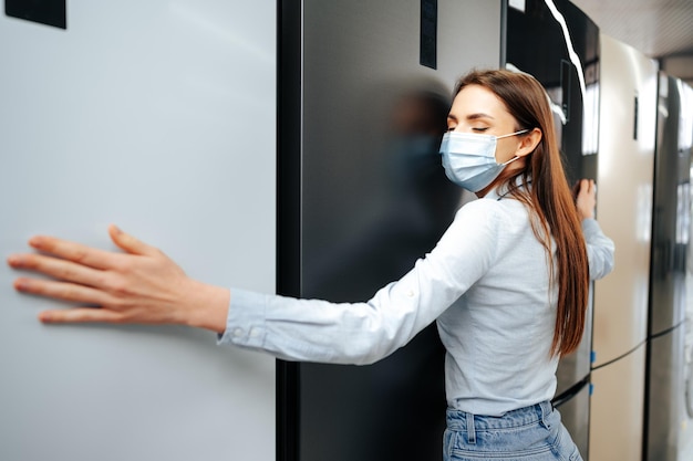 Young happy woman in mask leaning on her new refrigerator in a mall