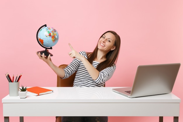 Young happy woman pointing on globe, planning vacation while sit, work at office at white desk with contemporary pc laptop isolated on pastel pink background. Achievement business career. Copy space.