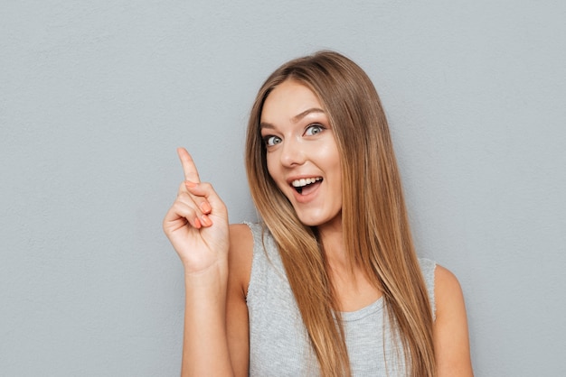 Young happy woman poiting finger up at copyspace isolated on a gray background