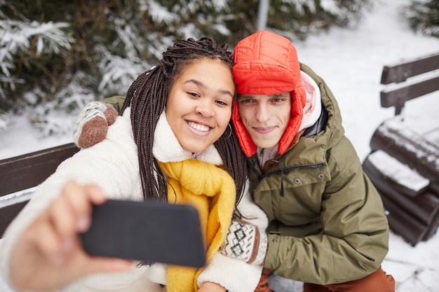 Young happy woman posing at camera of her mobile phone together with man while they sitting on bench in winter