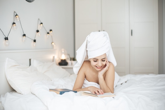 Young happy woman reading paper book in bed. Female person with towel on head rest in bedroon with book.