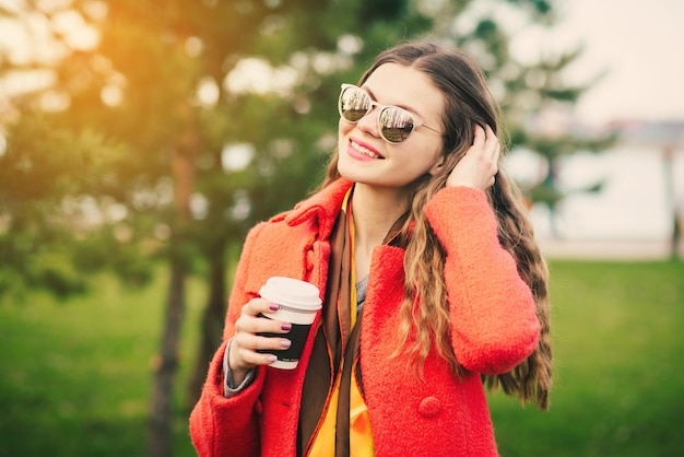 Young happy woman in red coat with coffee walking near trees in the park