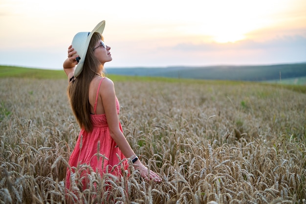 Young happy woman in red summer dress and white straw hat standing on yellow farm field with ripe golden wheat enjoying warm evening.