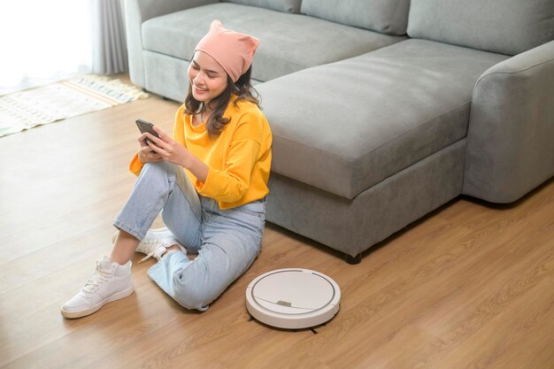 Young happy woman relaxing and using smartphone in living room while Robotic vacuum cleaner working