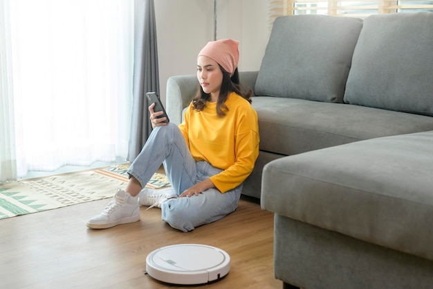 Young happy woman relaxing and using smartphone in living room while Robotic vacuum cleaner working