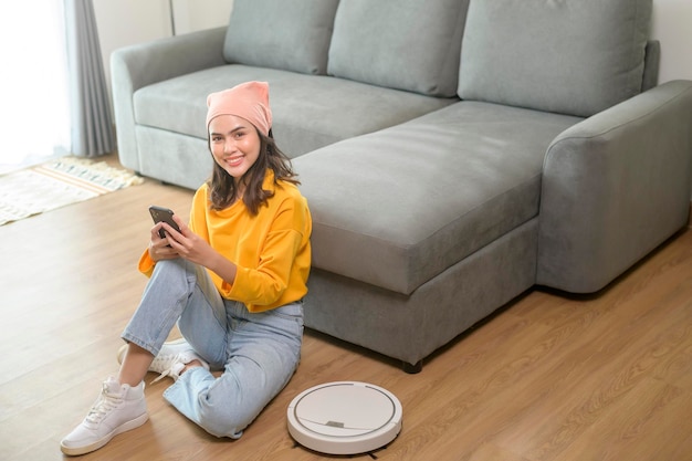 Young happy woman relaxing and using smartphone in living room while Robotic vacuum cleaner working