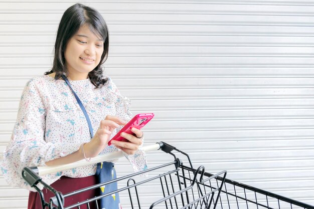 Young happy woman shopping cart Prepare to go shopping Are using a smartphone to search for products Promotional products and price discounts The background is a white steel door