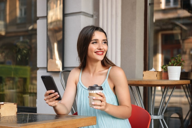 young happy woman sitting in cafe outdoors while using mobile phone and drinking coffee