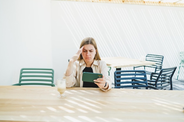 Young happy woman sitting at outdoor cafe table and talking on phone with cup of coffee smiling woman enjoying telecommuting in cafe or studying online on laptop