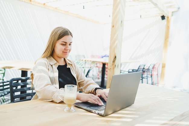 Young happy woman sitting at outdoor cafe table and talking on phone with cup of coffee smiling woman enjoying telecommuting in cafe or studying online on laptop