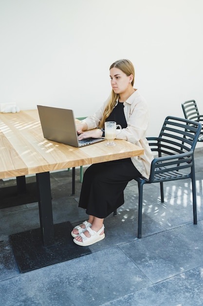 Young happy woman sitting at outdoor cafe table and talking on phone with cup of coffee smiling woman enjoying telecommuting in cafe or studying online on laptop