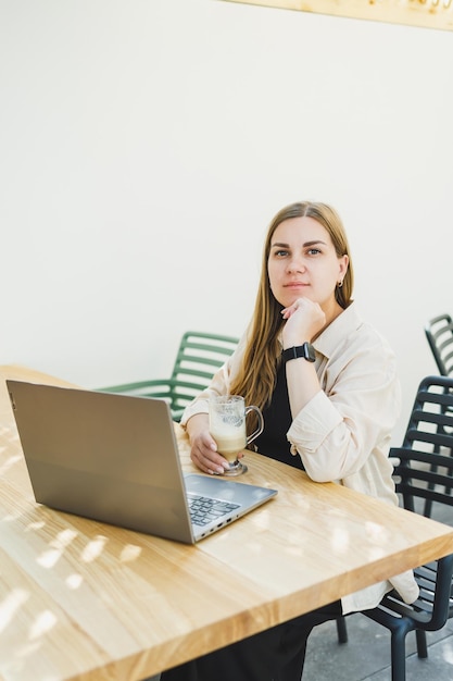 Young happy woman sitting at outdoor cafe table and talking on phone with cup of coffee smiling woman enjoying telecommuting in cafe or studying online on laptop