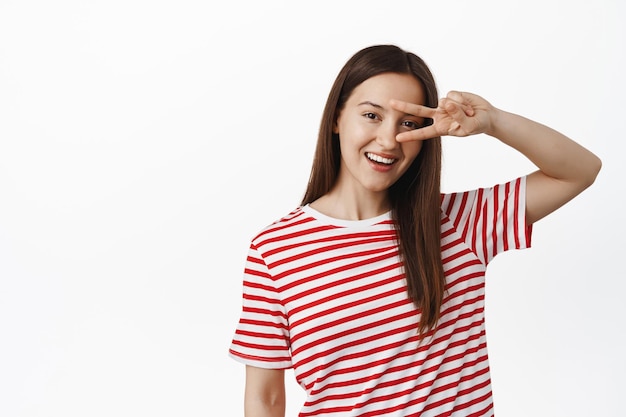 Young happy woman stay positive, enjoy summer, showing peace v-sign near one eye and smiling white teeth, standing in red t-shirt against white background.