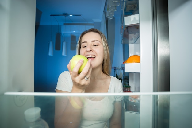 Young happy woman taking green apple out of refrigerator