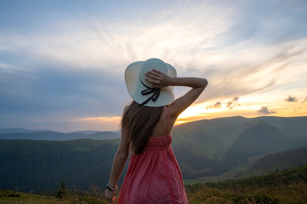 Young happy woman traveler in red dress standing on grassy hillside on a windy evening in summer mountains enjoying view of nature at sunset.